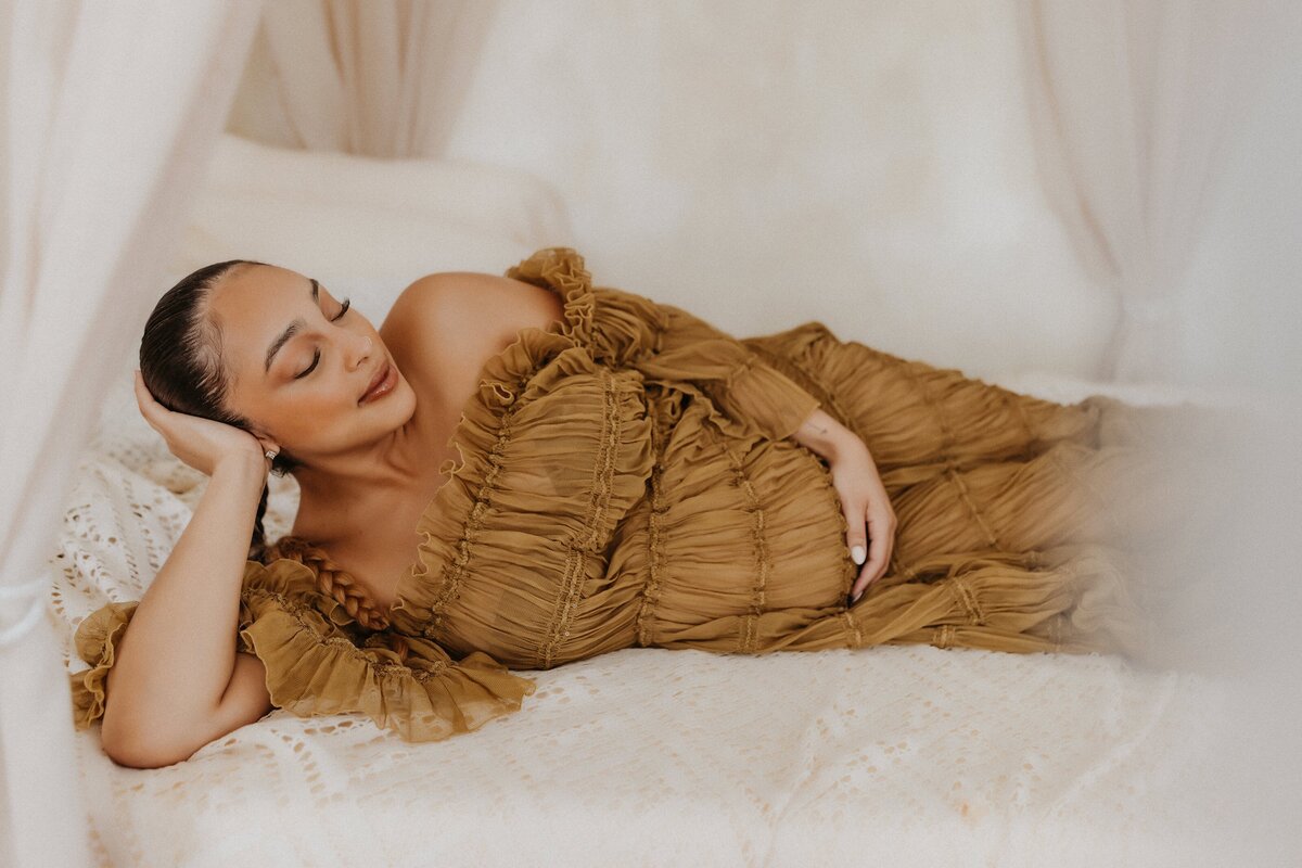 A person, perhaps captured in elegant Macon maternity photography, lies on a bed wearing a ruffled brown dress. One hand rests under their head and the other on their stomach. Their eyes are closed, surrounded by light-colored curtains that softly frame the serene scene.