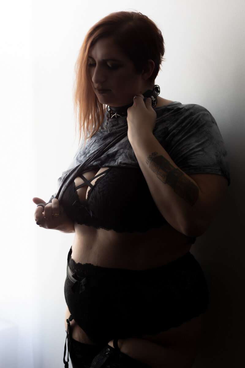 a plus size woman with tattoos poses for marquise photography in strappy lingerie and an alternative crop top that she is pulling off of her.