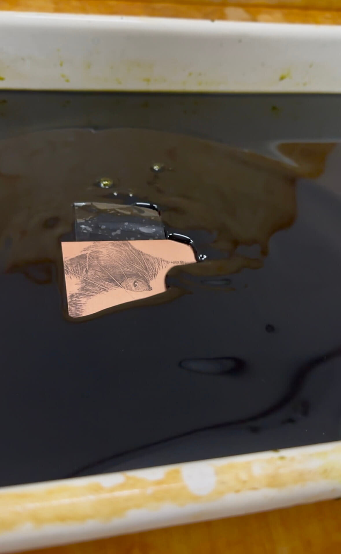 A copper plate partially submerged in an etching bath, revealing an image developed from a toner transfer.
