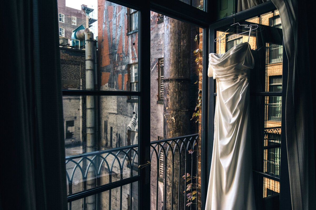 A white wedding dress hangs on a balcony door with an urban, industrial scene in the background..  This image was taken at a Greenwich Hotel wedding