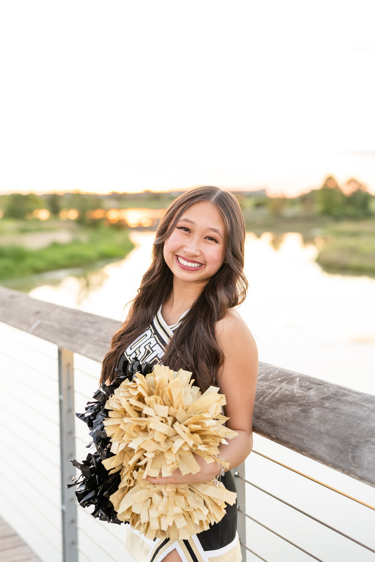 Houston High School senior girl in cheerleading outfit smiling while holding poms and leaning against bridge at Josey Lake Park