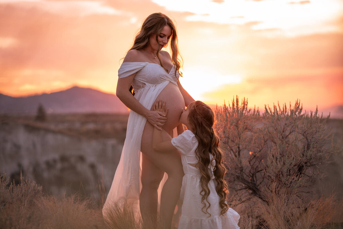 A pregnant women stands against a dramatic sunset and her young daughter crouches down to kiss her exposed belly. Image by Brittany Danielle Photography