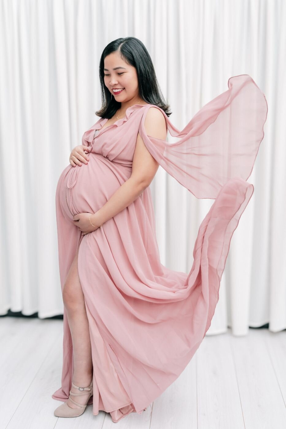 Pregnant woman in flowing blush-pink gown smiling and holding her belly as the gown’s fabric streams gently behind her.