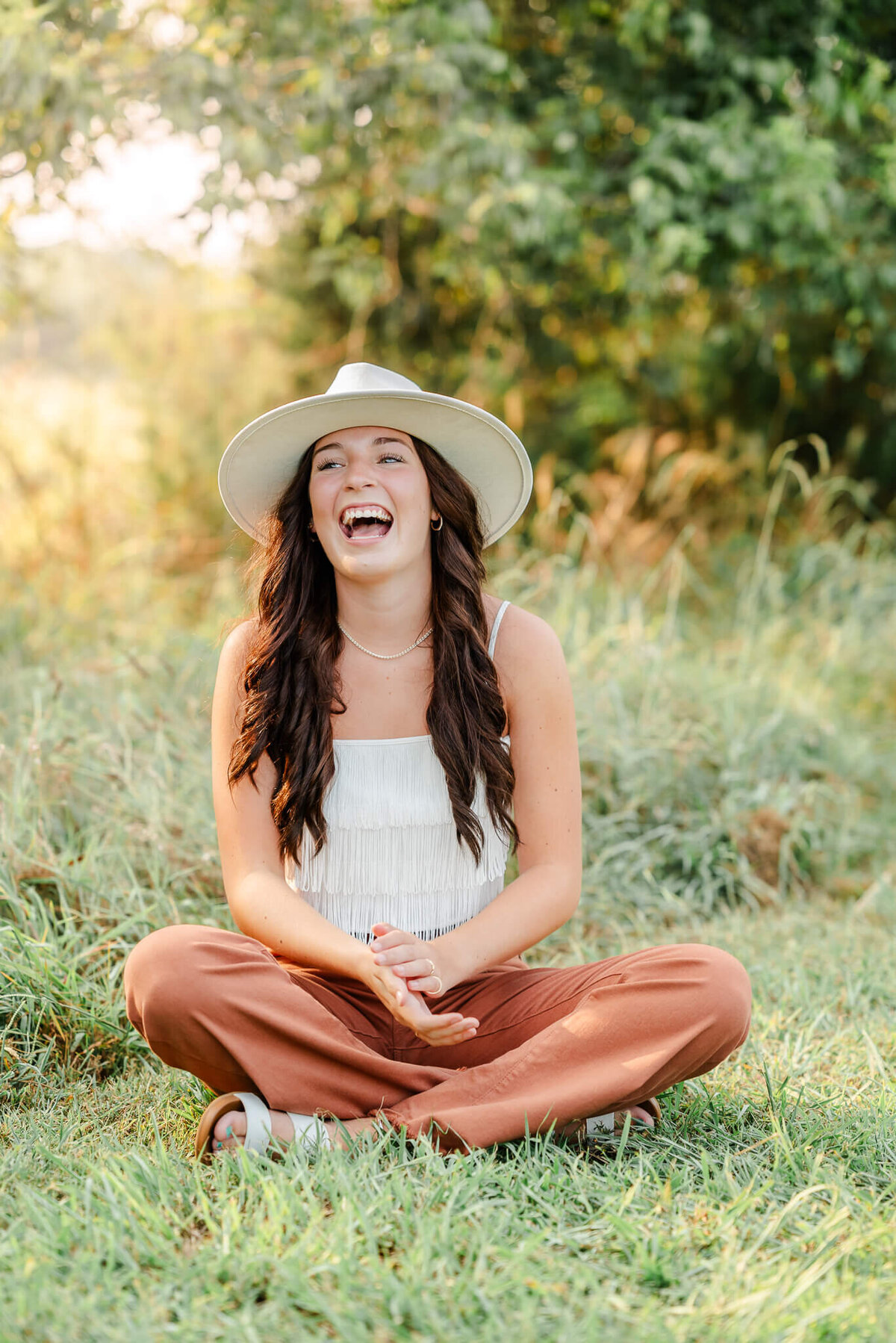 A high school senior wearing orange pants, a white fringe top, and a white hat sits in the grass and laughs. Photo taken at Bells Mill Park in Chesapeake by Justine Renee Photography.