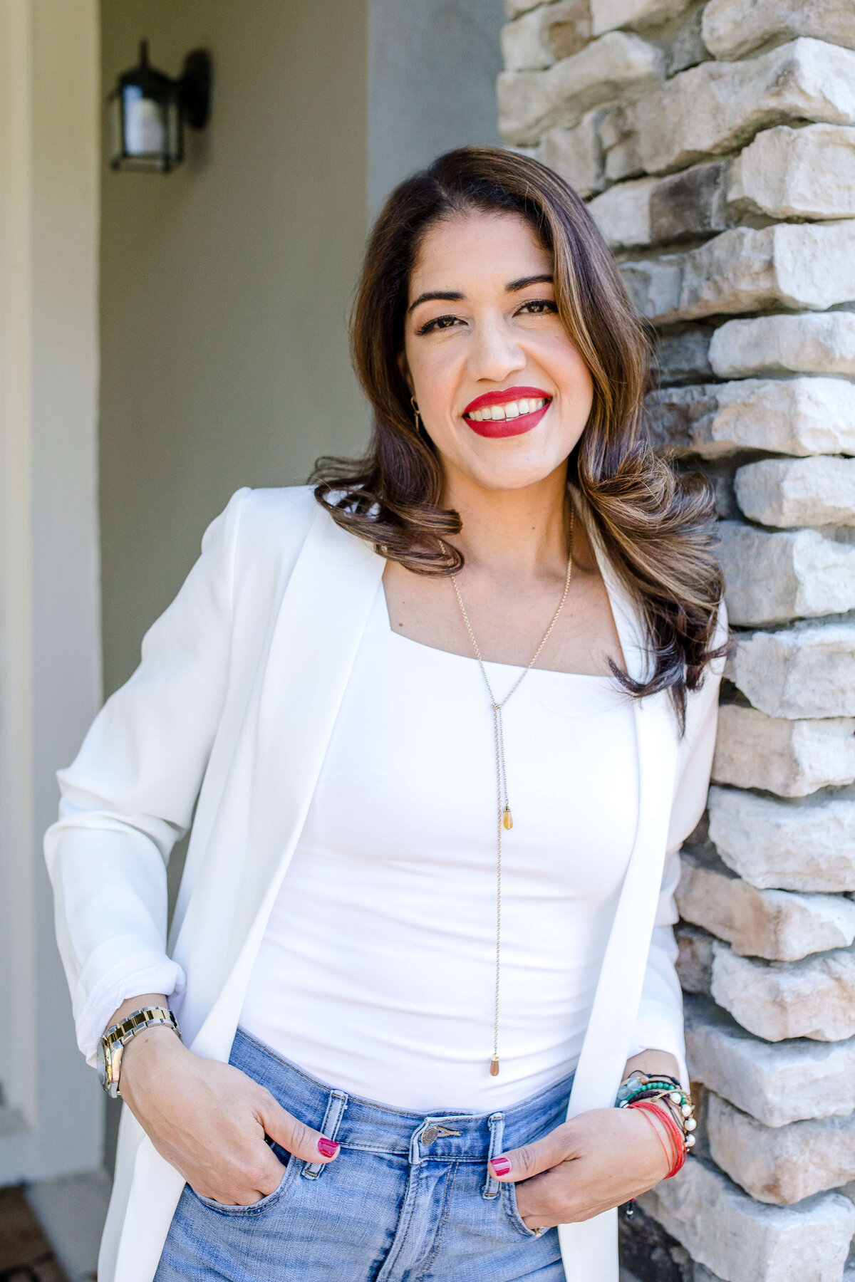 Woman in white shirt and blazer with red lipstick on hold her hands in her Jean pockets and smiles while leaning against a stone pillar for her branding photo shoot with brand photographer near me