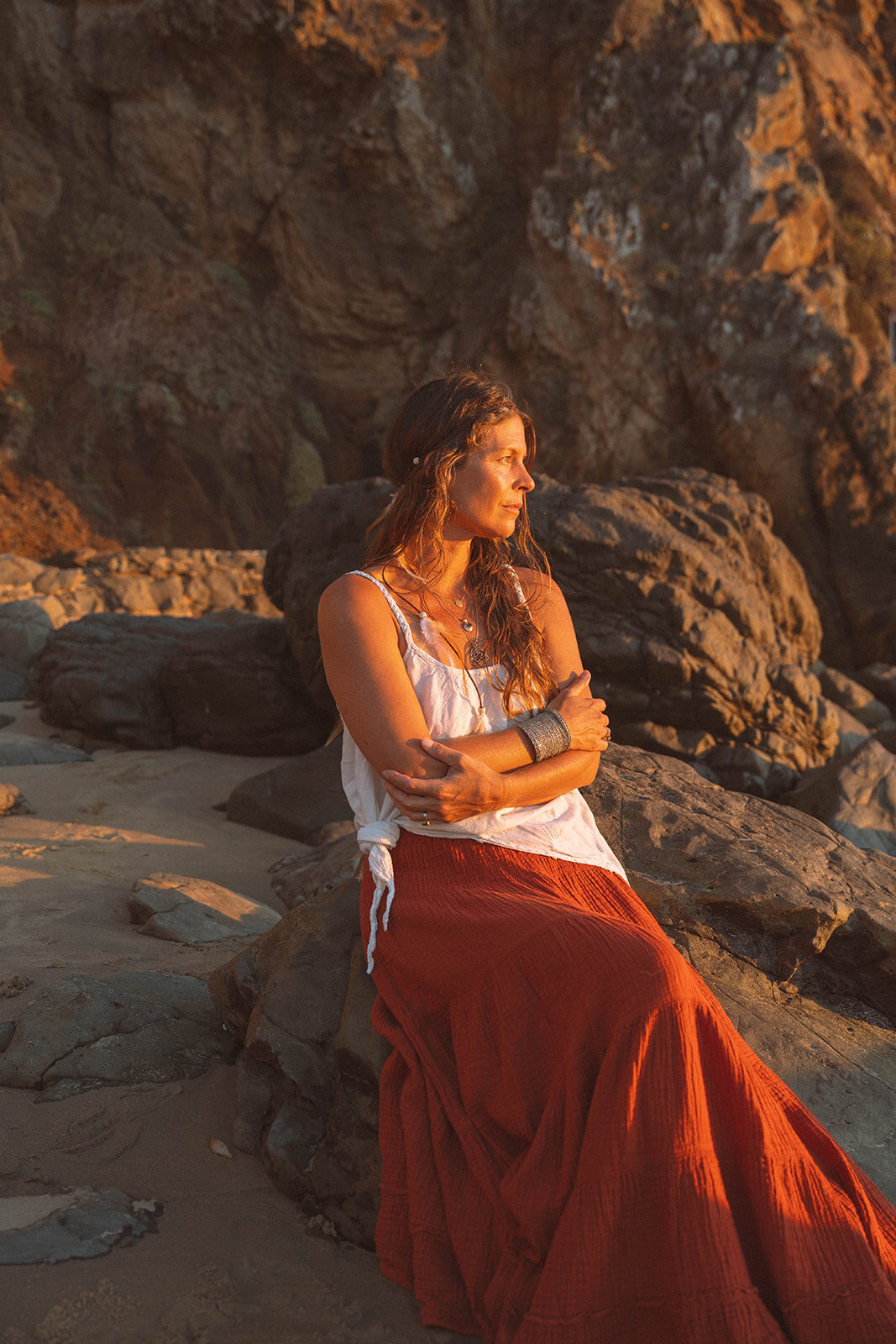 Woman sitting on coastal rocks at sunset during a branding session in Laguna Beach, California. Soft golden light highlights natural textures and warm tones.