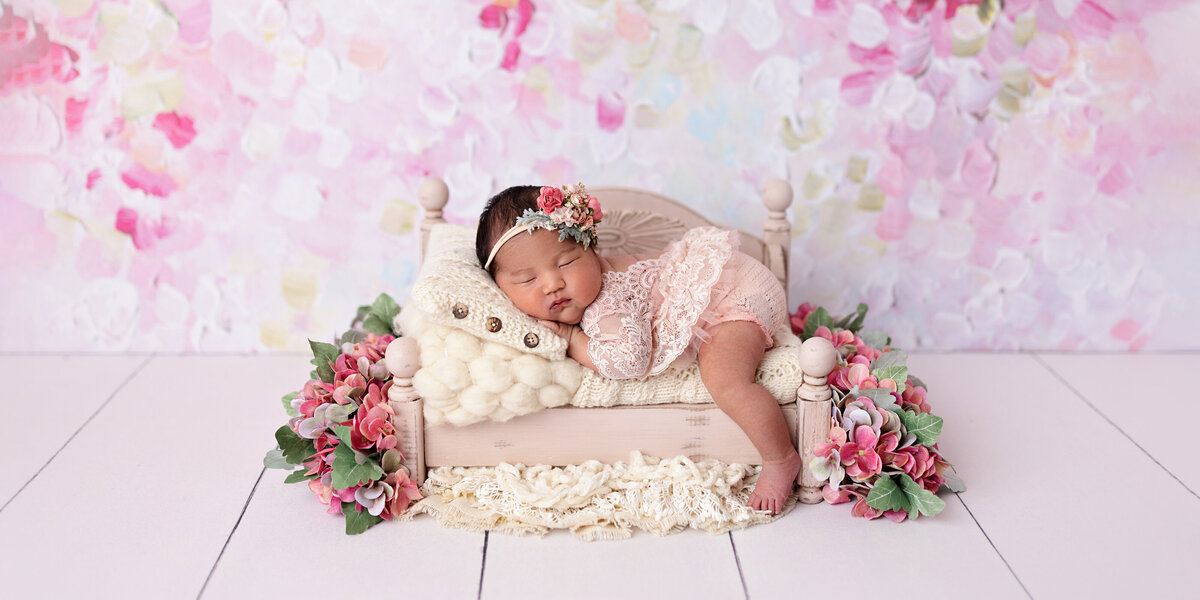 Newborn baby girl sleeping on a miniature cream bed surrounded by pink hydrangeas and textured blankets against a pastel floral backdrop.