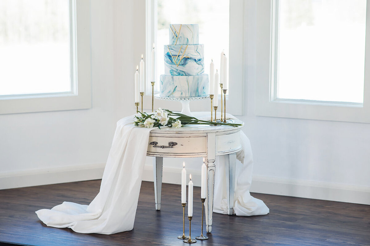 3 tiered blue and white  marbled wedding cake with gold geometric details, surrounded by white candles and greenery, by Yvonne's Delightful Cakes, classic cakes & desserts in Calgary, Alberta, featured on the Brontë Bride Vendor Guide.
