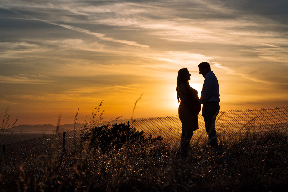 silhouette of maternity couple standing on hill at sunset in Oakland