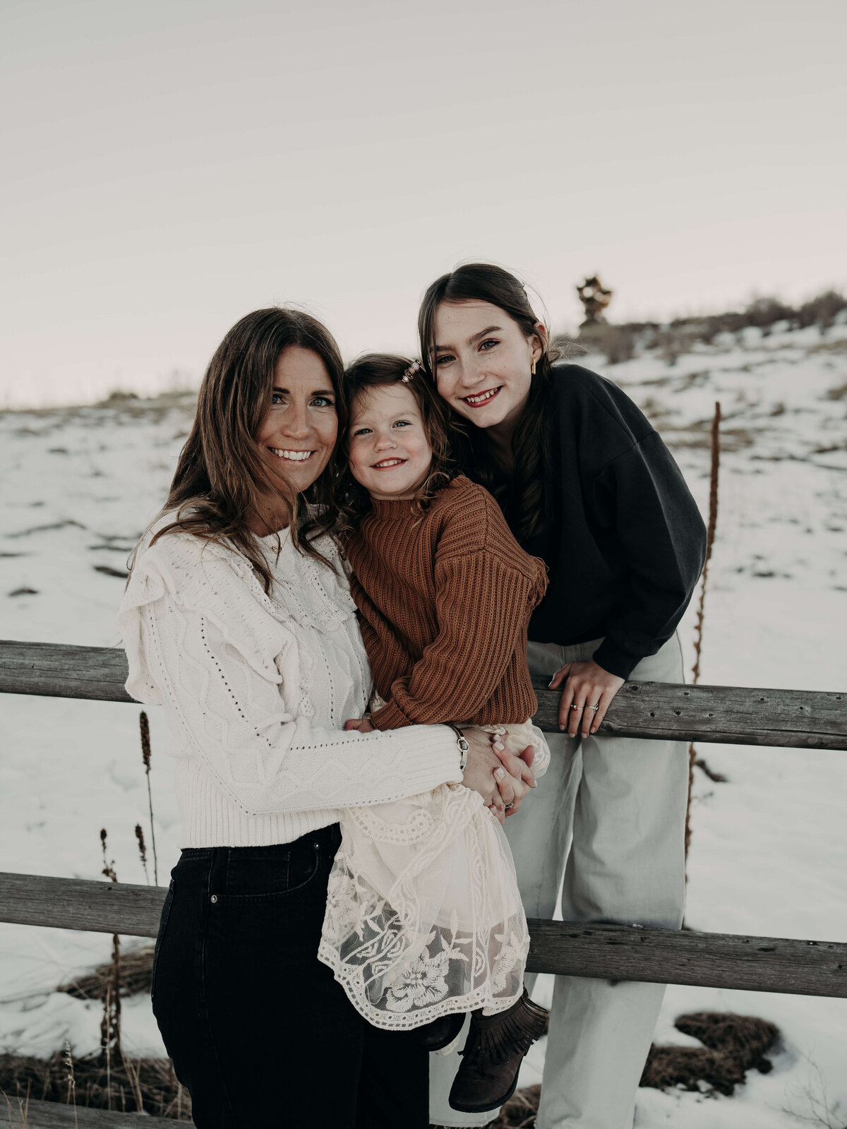 A portrait of a mother with her two daughters during an outdoor winter photography session. All three girls were coordinating color, pallets of Browns and creams as they smile at the camera.