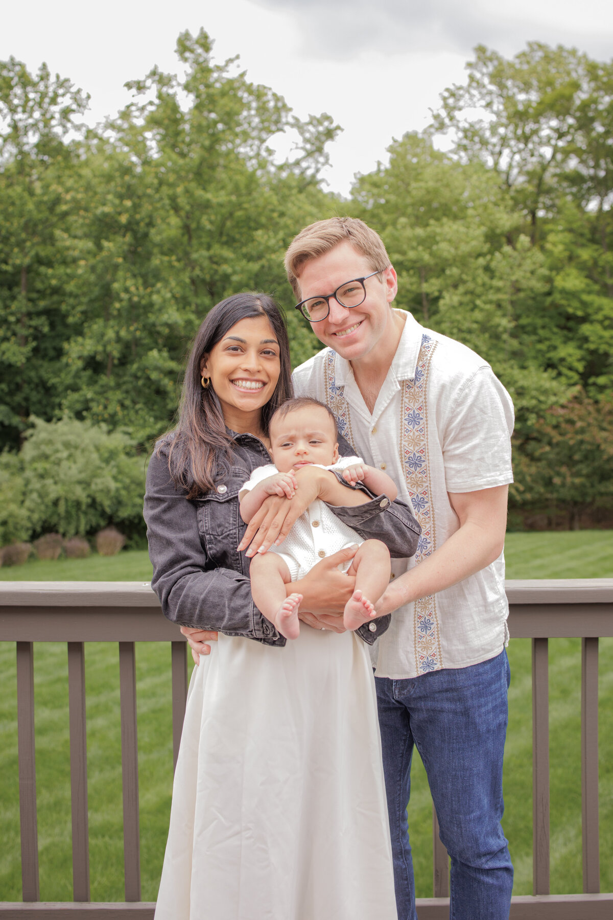 Smiling parents holding their baby during an outdoor family photo session on a deck surrounded by greenery.