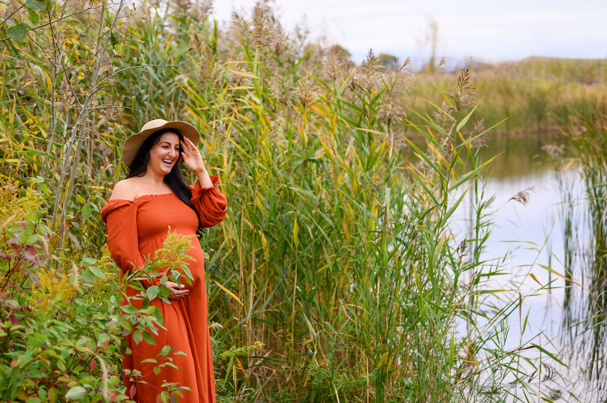 pregnant woman wearing an orange maternity dress and beige hat with a pond in the background for her fall outdoor maternity photos