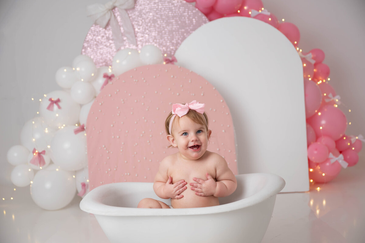 Smiling baby sitting in a mini tub, wearing a pink bow, with pink and white balloon and arches in the background.”