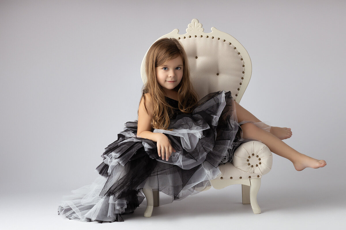 Dallas family photographer captures a young girl in a black and white ruffled gown, sitting elegantly on a vintage chair.