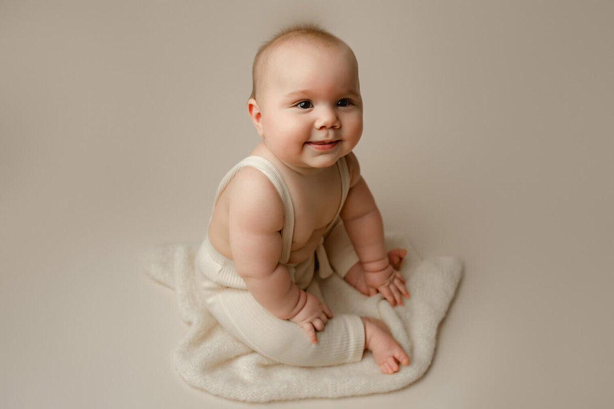 A baby boy sits on a white cloth with white suspenders on