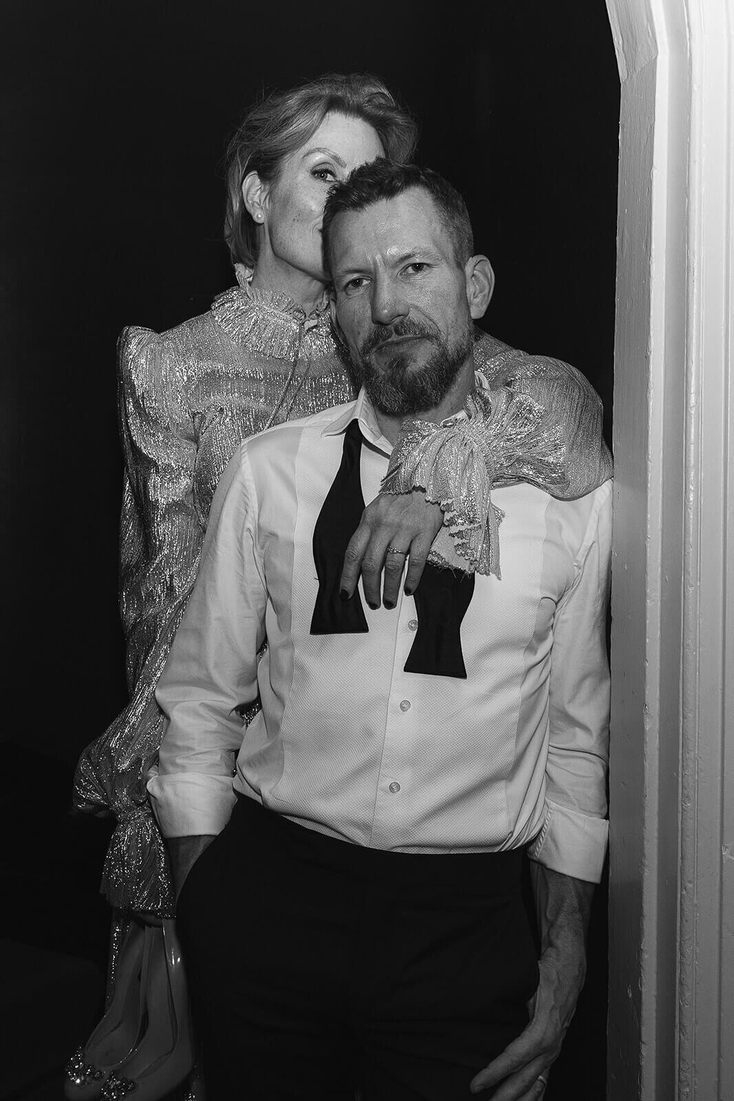 A bride hides her face and kisses the back of her grooms head. Her arm over his shoulder as he leans on a door frame. Bow tie undone.