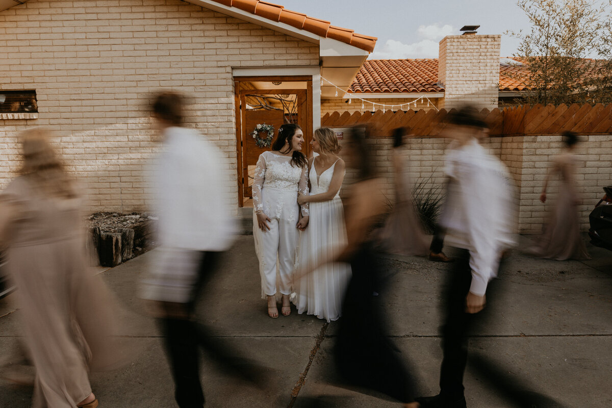 two brides standing together with their wedding party walking around them