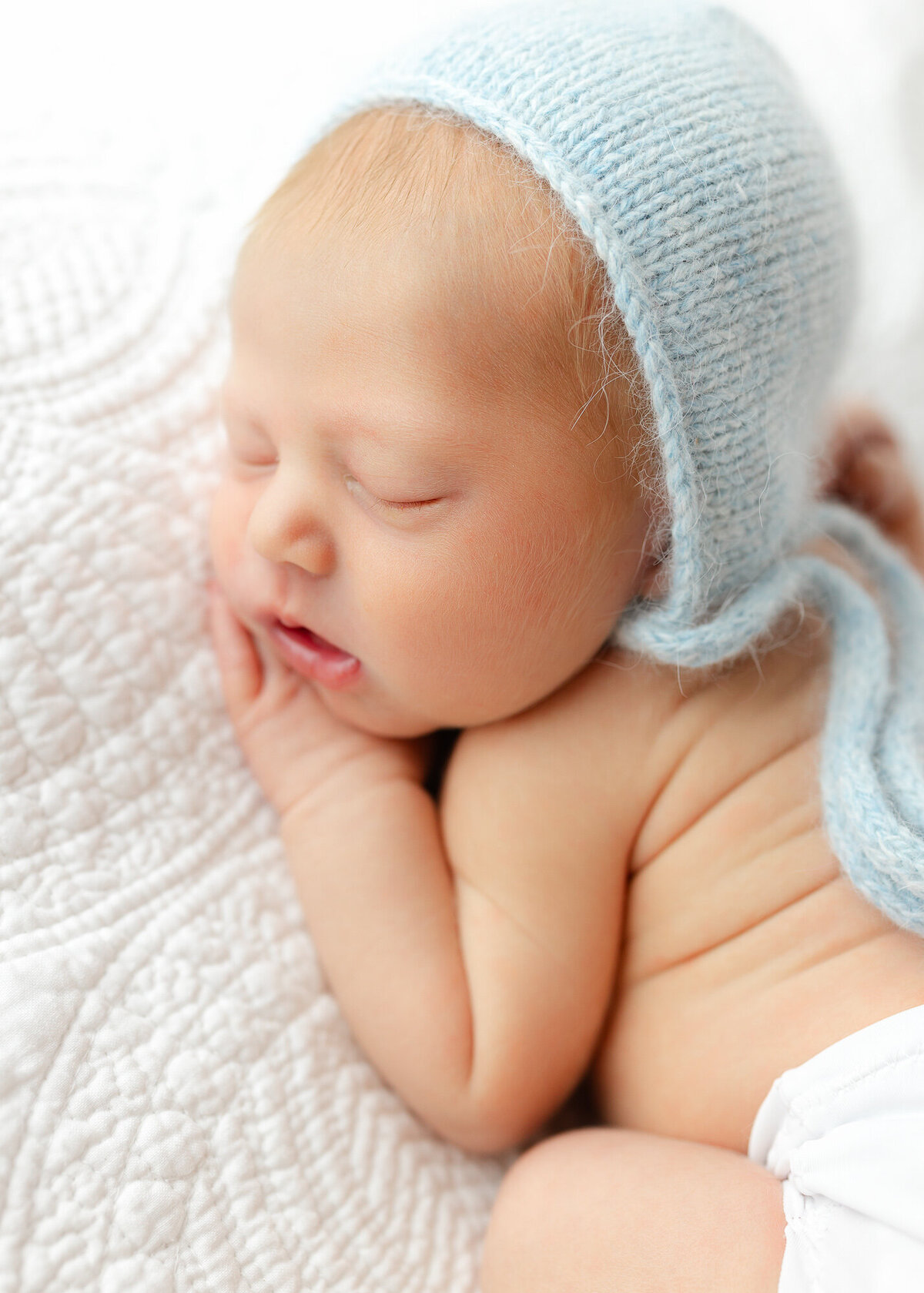 Newborn boy wearing a blue bonnet laying on a white embroidery blanket