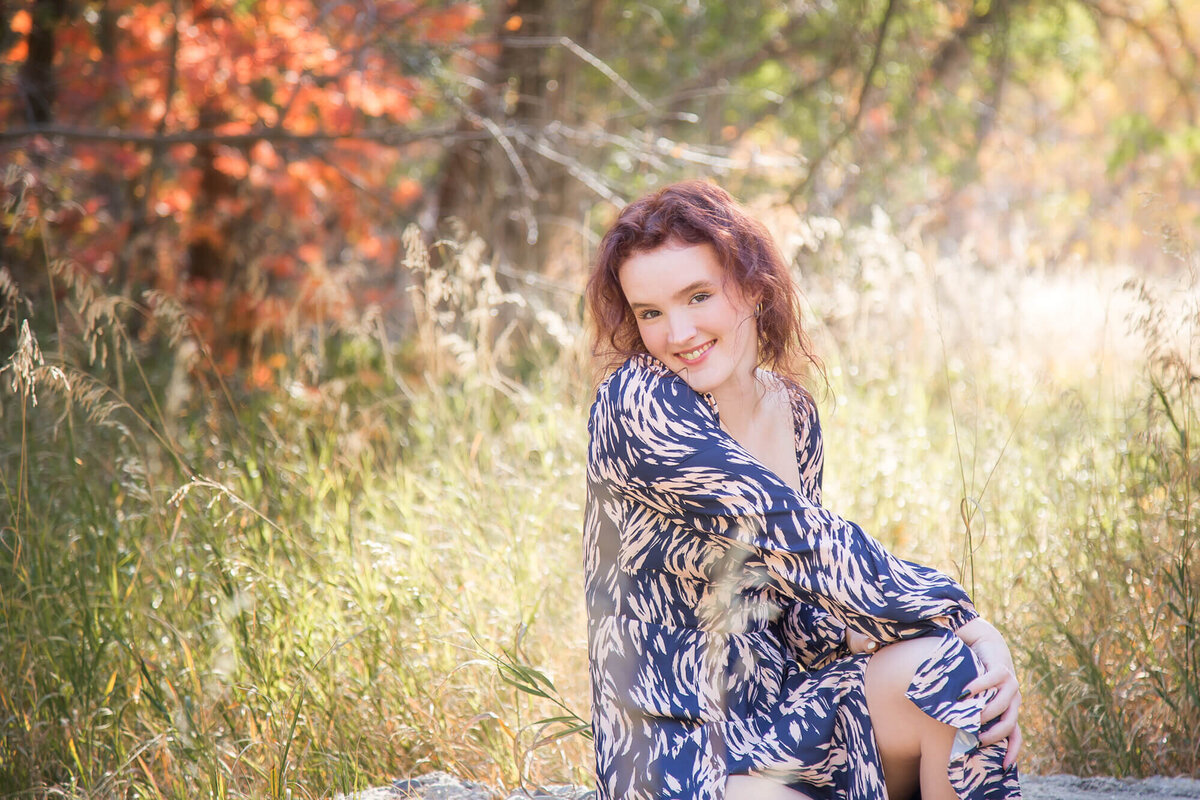 redheaded young woman shyly smiling while sitting on a log in a field of grass