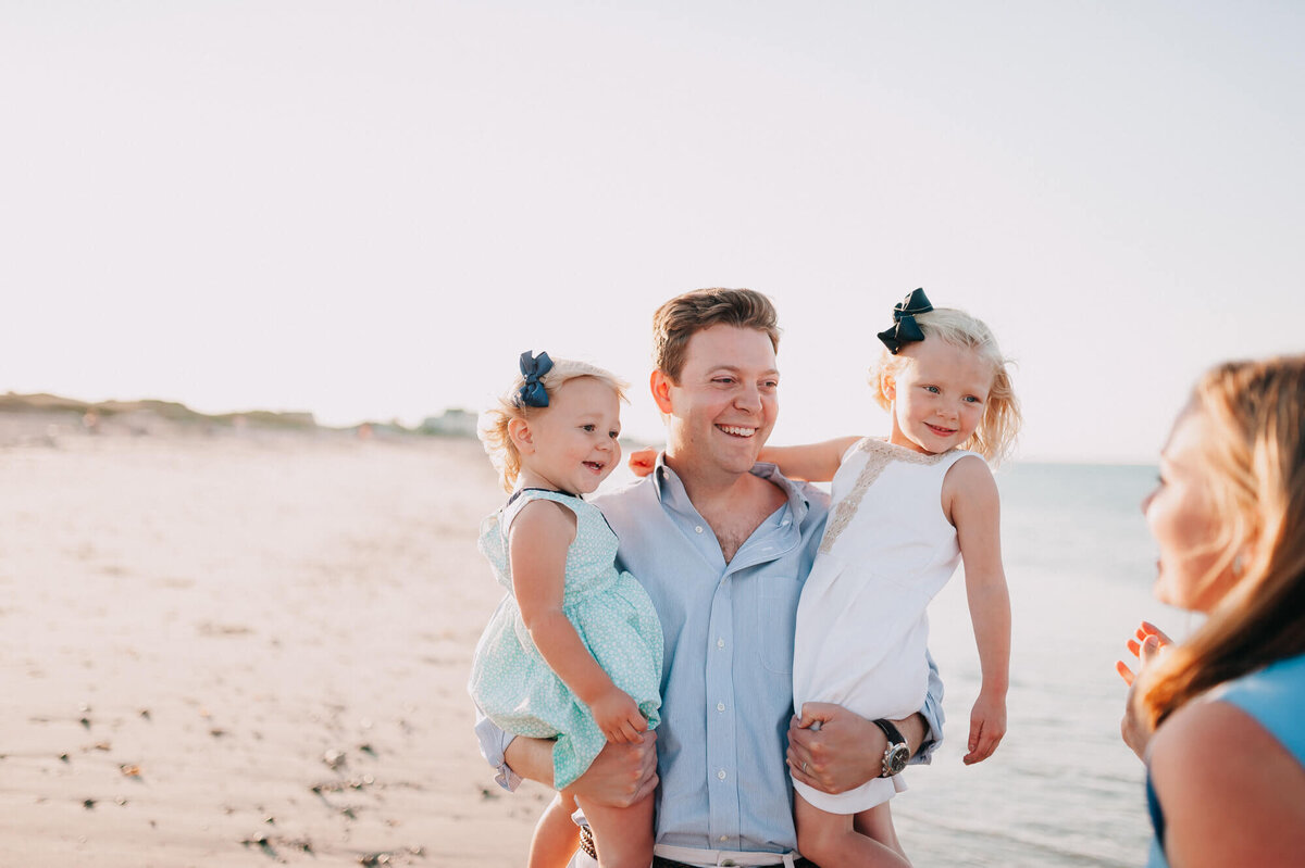 A father holds his two young daughters on the beach, they are all smiling and looking at the girls' mother
