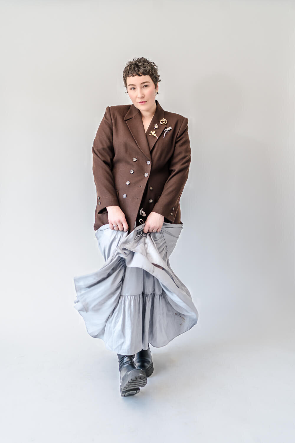 Woman in structured brown blazer and tiered silver dress walking toward camera in studio.