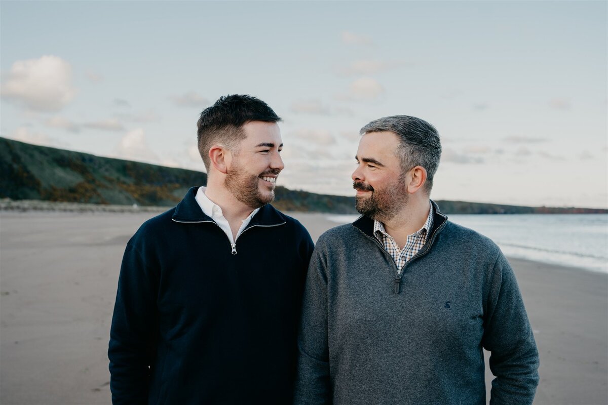 An LGBTQ+ couple stand on a beach and look at each other during their Aberdeen pre-wedding photography session.