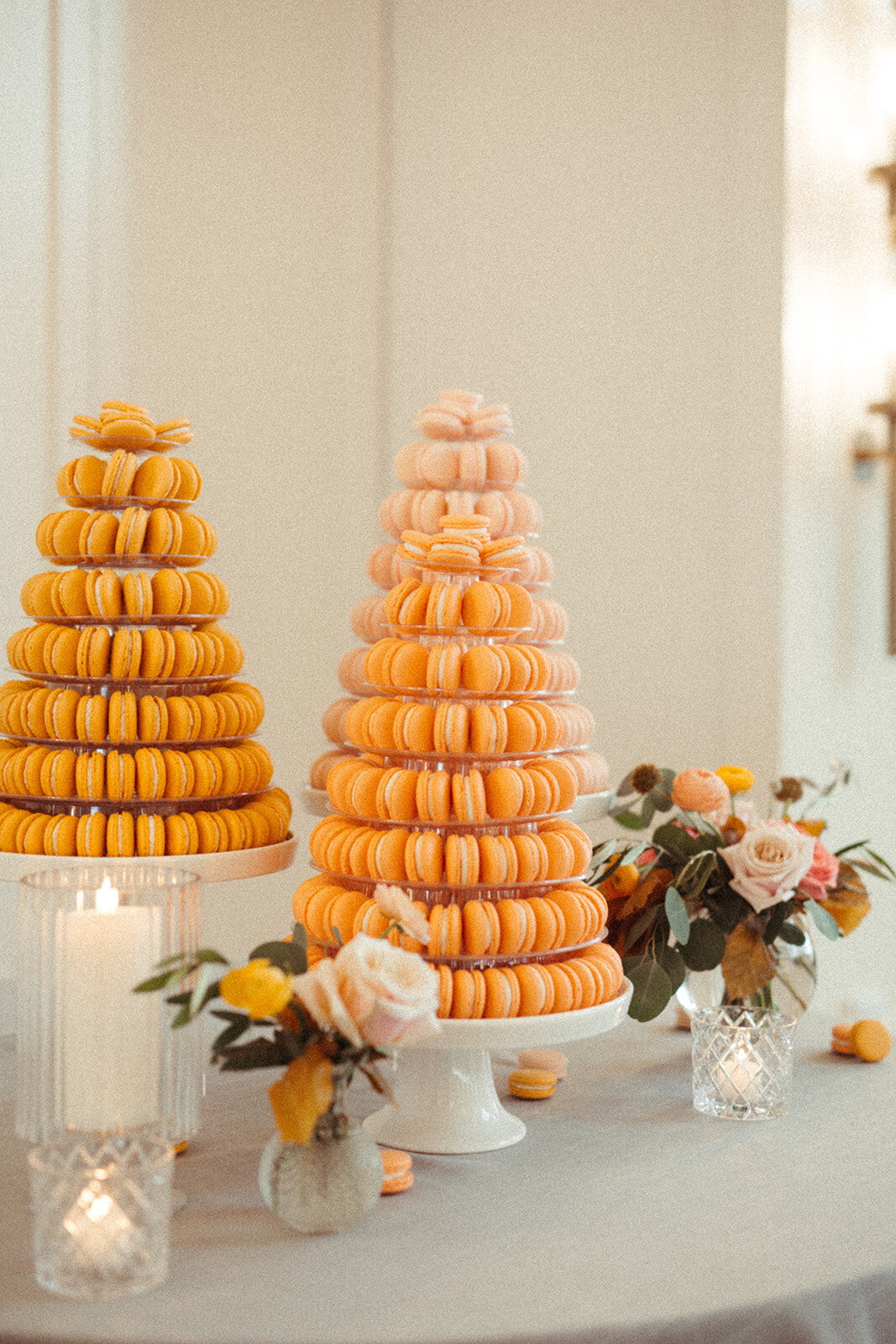 Two peach and orange macaroon towers on cake stands atop a table with candlesticks and flowers.