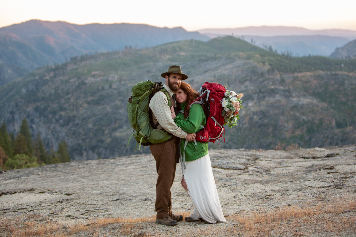 A bride and groom stand holding each other as the bride rests her head on his chest. They are both wearing backpacks and there are mountains behind them.