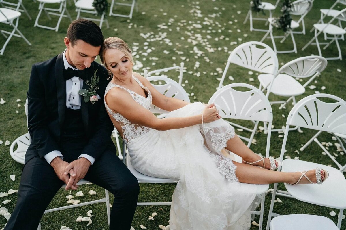 A bride and groom sitting on white chairs on a lawn sprinkled with rose petals, sharing a quiet and intimate moment post-ceremony.
