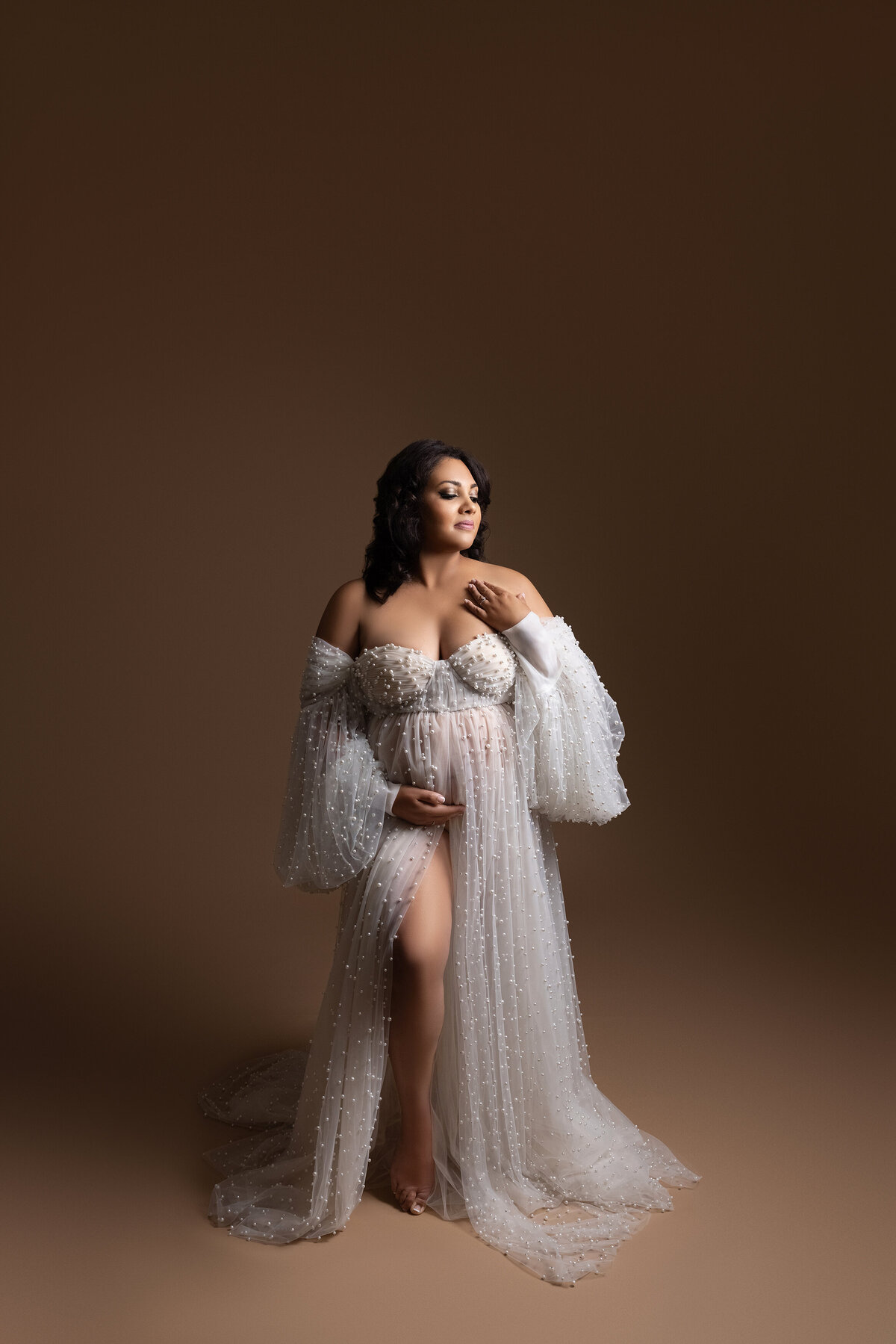 Maternity photos captured by top London, ON Maternity Photographer Amy Perrin-Ogg.  Expectant mom in elaborately beaded cream coloured gown is standing facing the camera with one hand touching her shoulder and the other hand under her bump. Her eyes are closed and head positioned toward soft light.