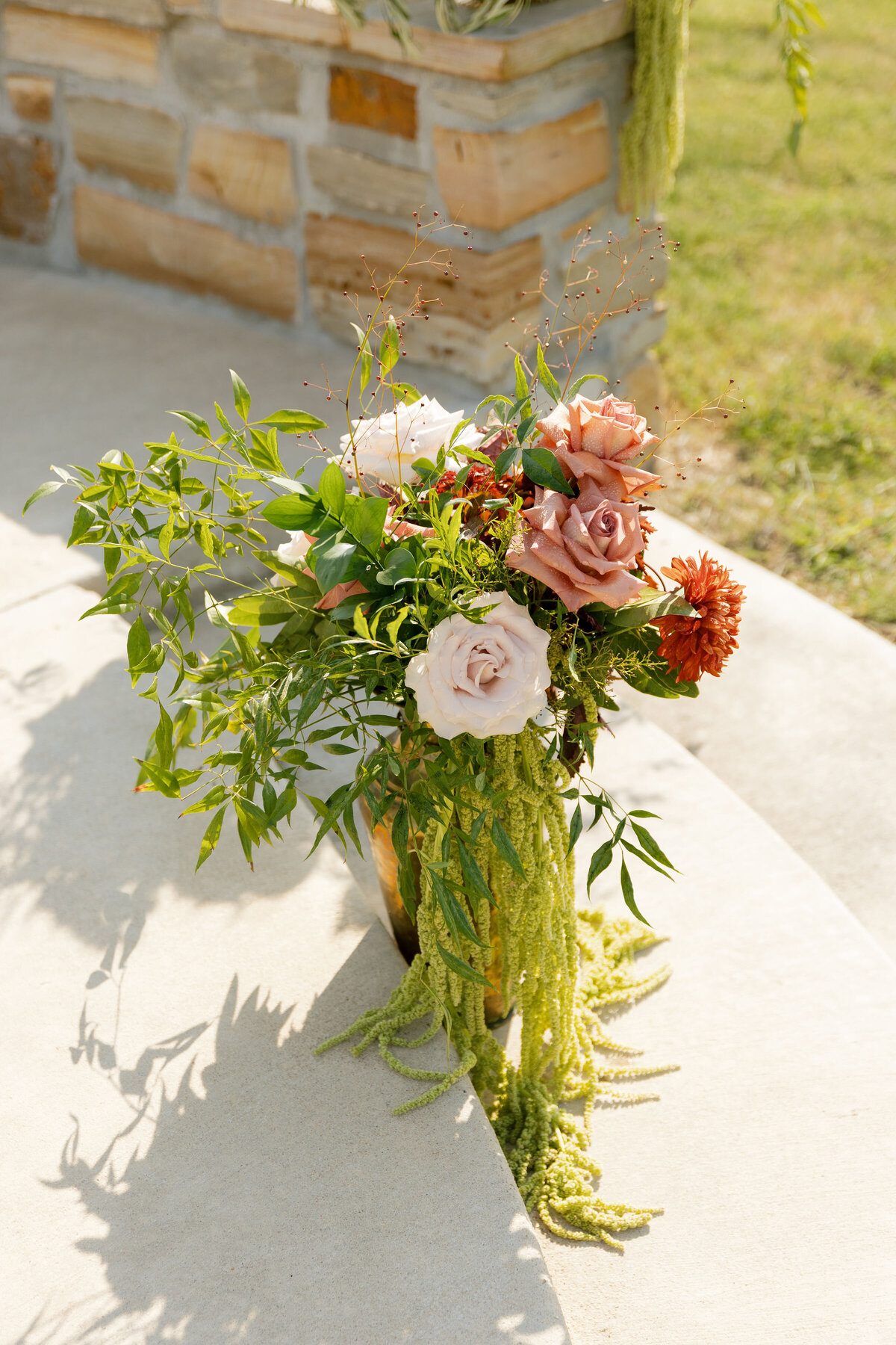 A detailed close-up of a wedding floral arrangement highlighting large blush roses, peach roses, rust-colored blooms, and abundant greenery spilling over the vase with long, trailing amaranthus. The arrangement sits on light concrete with soft shadows, adding organic movement and texture to the outdoor ceremony décor.