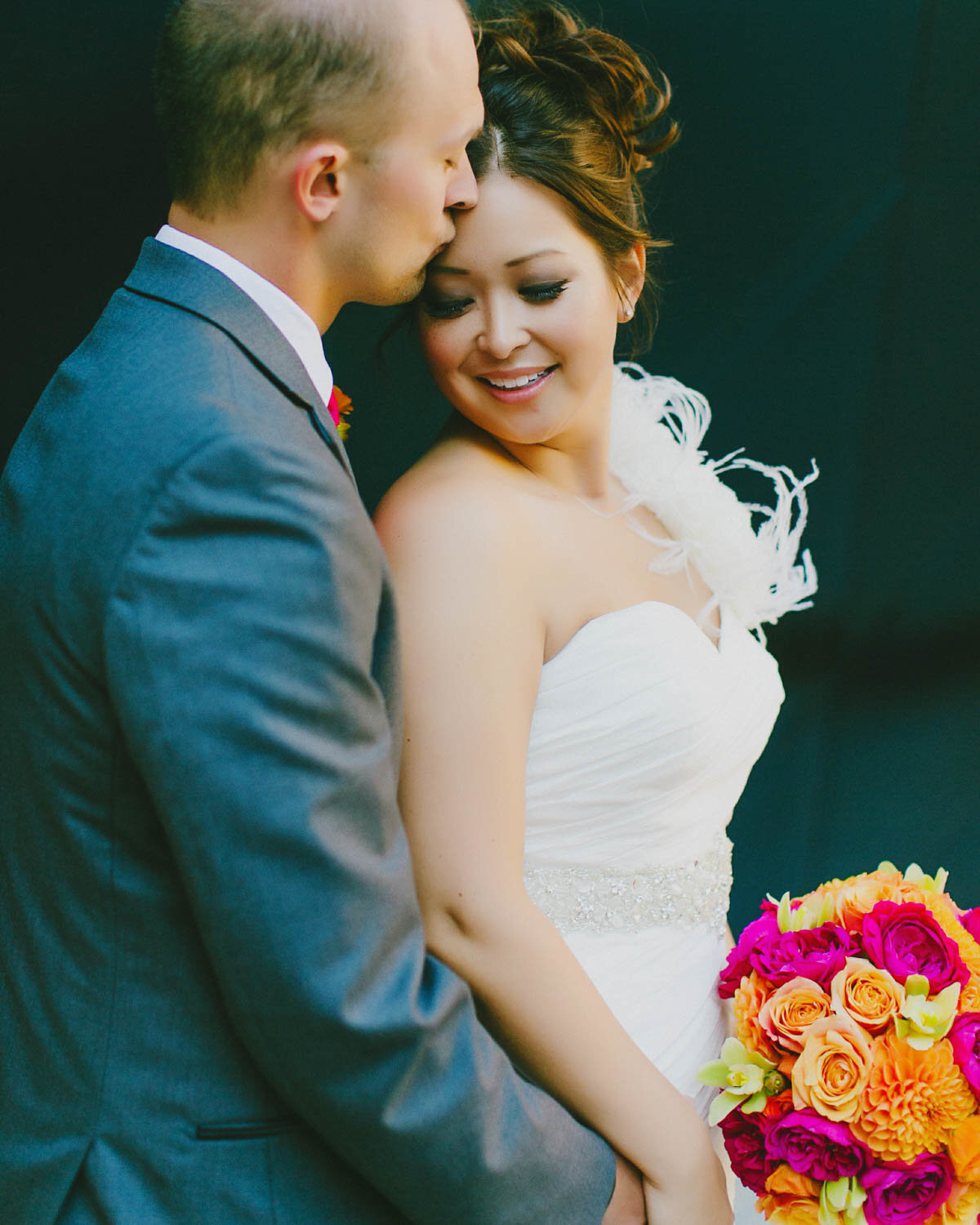Groom stands behind Bride kissing her forehead as she turns to her left as she smiles looking down