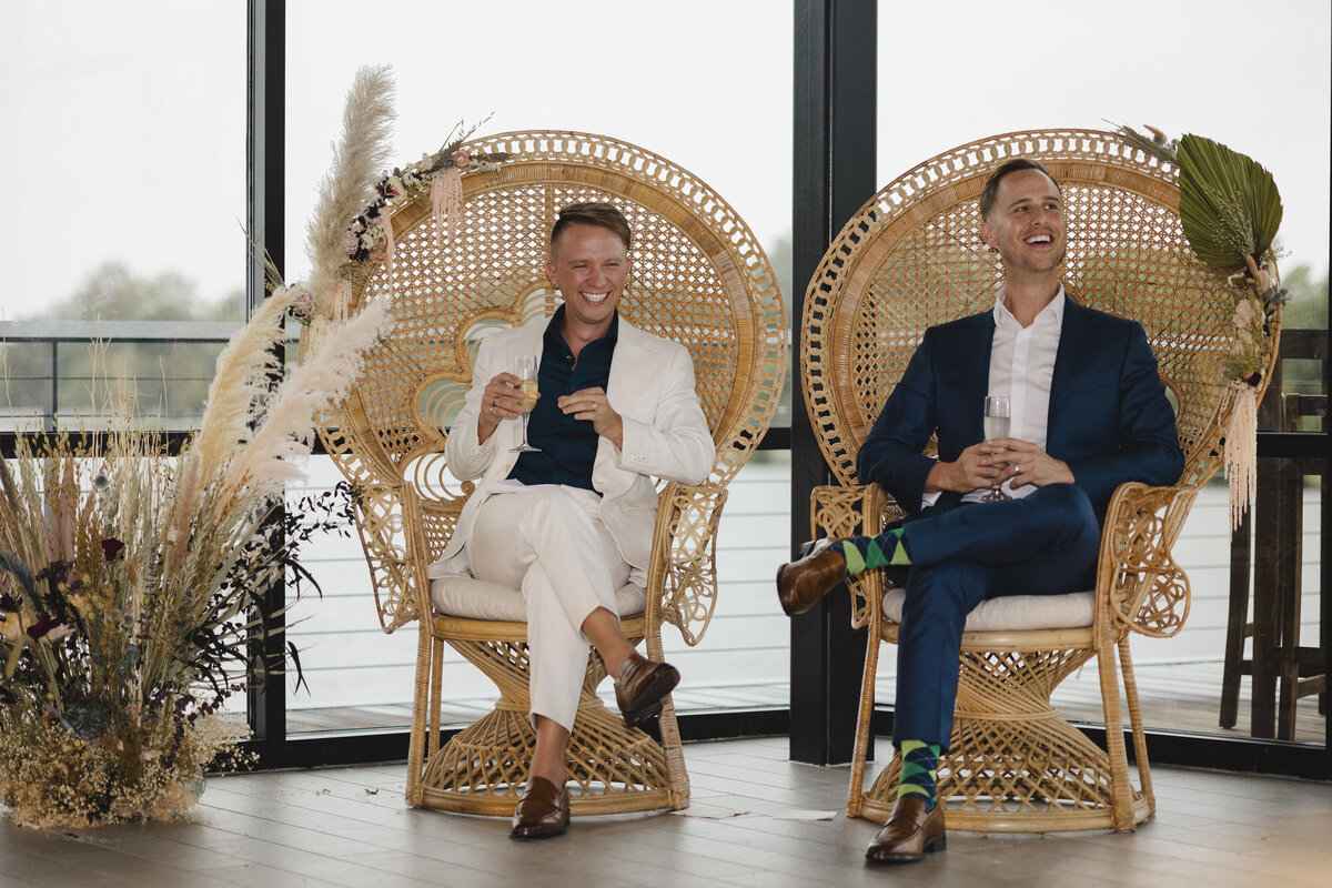 Two grooms sitting in wooden, woven chairs smiling 