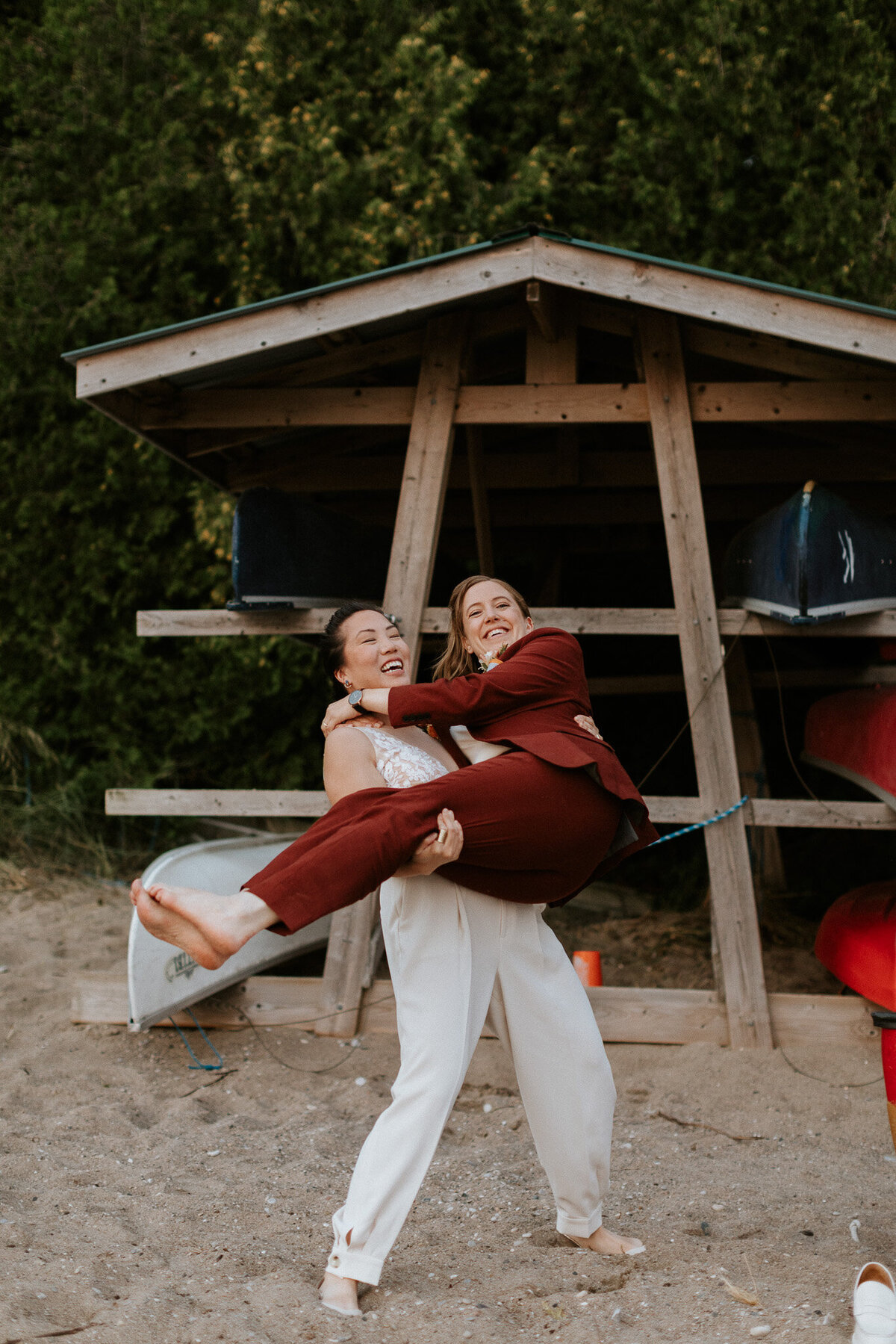 Two people are smiling, with one in a maroon suit playfully lifting the other in white attire in front of a wooden structure on a sandy beach, captured perfectly by a talented Toronto photographer.`