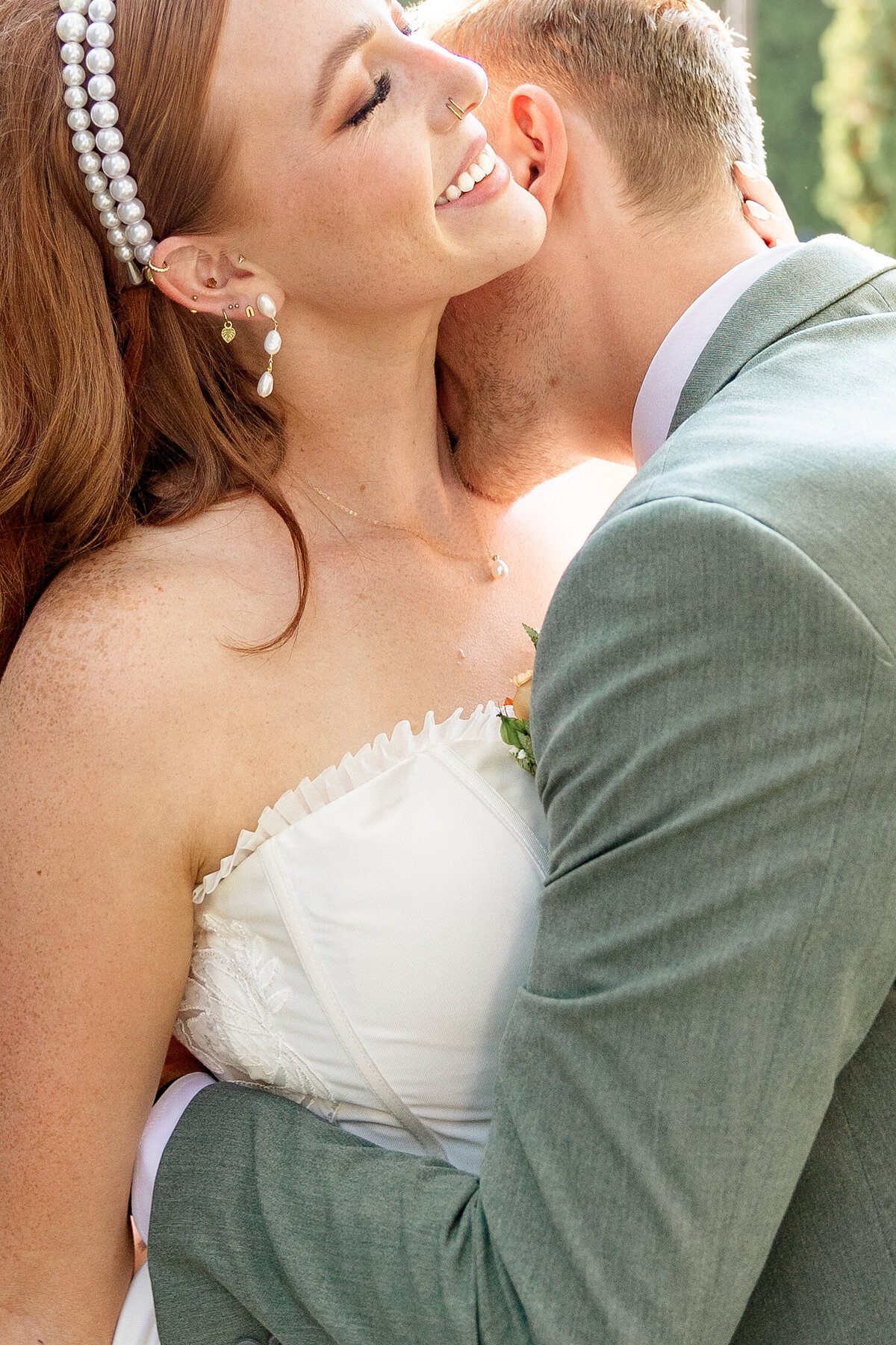 The groom wraps his arms around the bride and leans in to kiss her neck, as she smiles with joy in the sunlit garden.