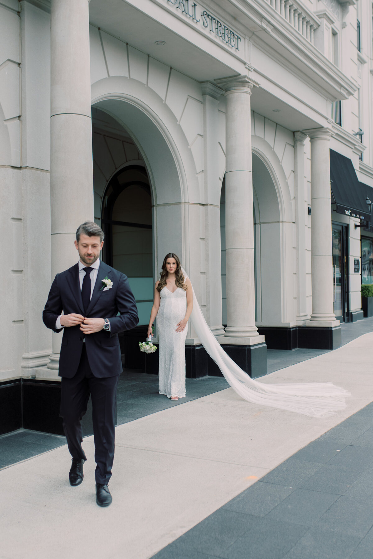Groom fixing suit as he is walking away from bride as bride poses on wall holding her stomach.