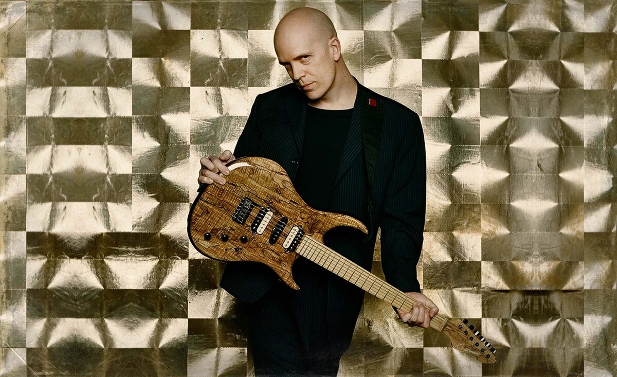 Male musician photo Devin Townsend wearing black suit while holding brown electric guitar against gold wallpaper