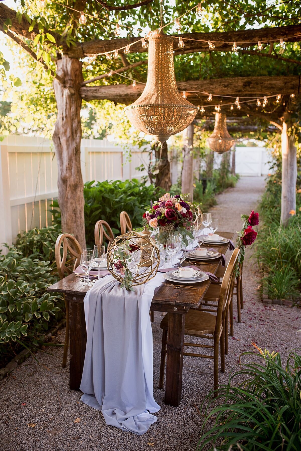 Elegant boho wedding under a rustic wood arbor with hanging wisteria  vines, string lighting and long brass filigree chandeliers. A dark wood farm table sits in a lush garden at Carnton Plantation. The farm table has a long soft gray organza table runner with wood chairs with geometric shaped backs. The table as large gold geometric spheres and a large floral centerpiece in a gold footed bowl. The flowers are burgundy dahlias, burgundy peonies, peach roses, blush tea roses, king protea, prince protea and greenery. Stacked plates of a white scalloped charger, a light gray dinner plate, a white salad plate with gold geometric patterns and a white bowl with gold flatware on either side of the plates.