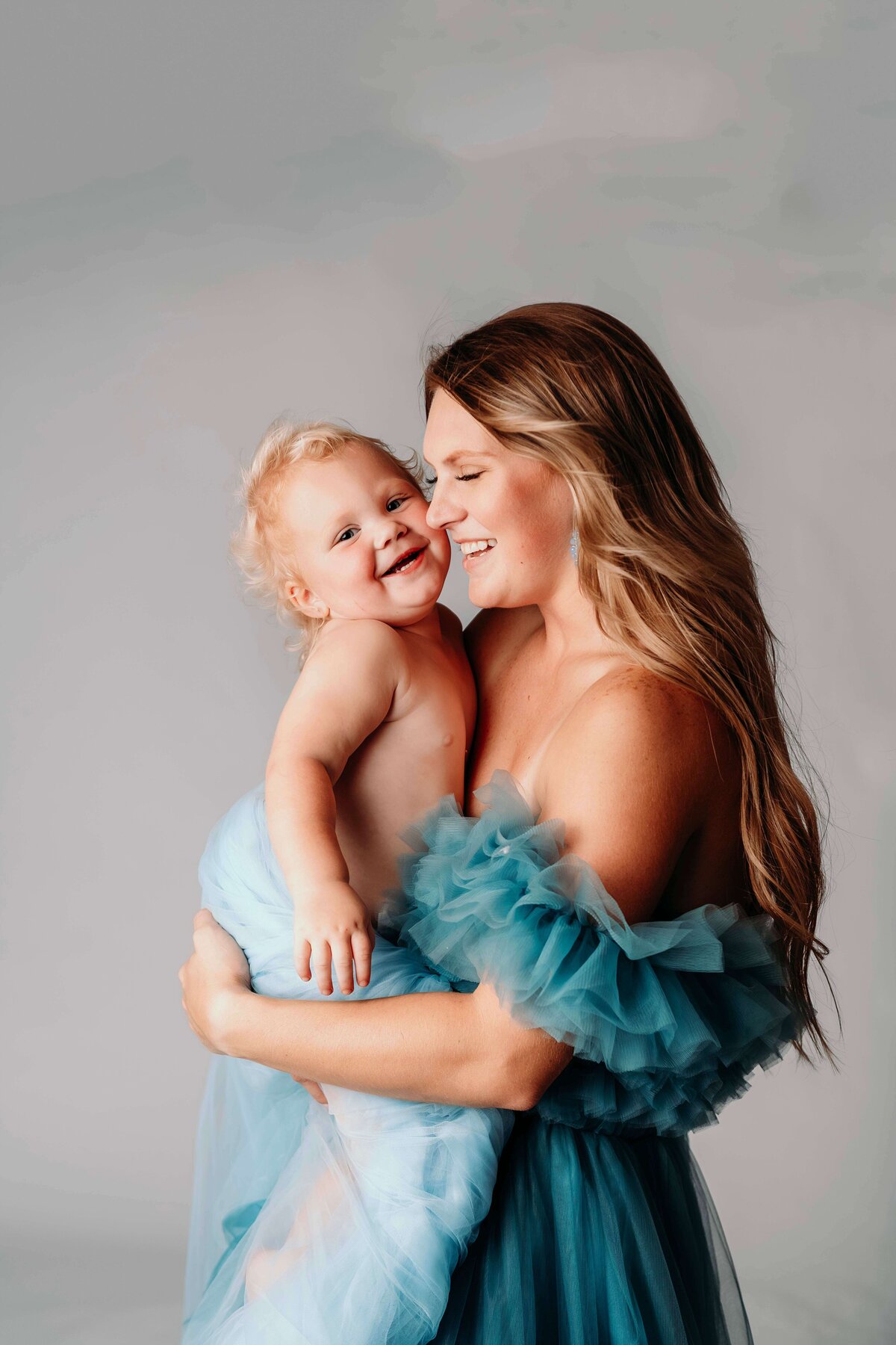 A close-up photo captures a young mom and her toddler daughter in matching blue tulle gowns. The child, looking very happy and playful, grins and waves at the camera. The mom, giggling at her daughter, holds her close and snuggles in for a warm, intimate moment, creating a tender motherhood portrait by Mobile, AL Motherhood Photographer