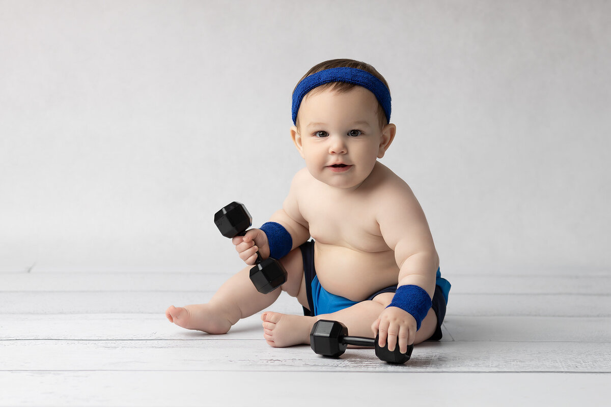 Baby boy dressed in blue wristbands, shorts, and a headband holds a pair of toy dumbbells while smiling on a white wood floor during a Dallas milestone photography session.