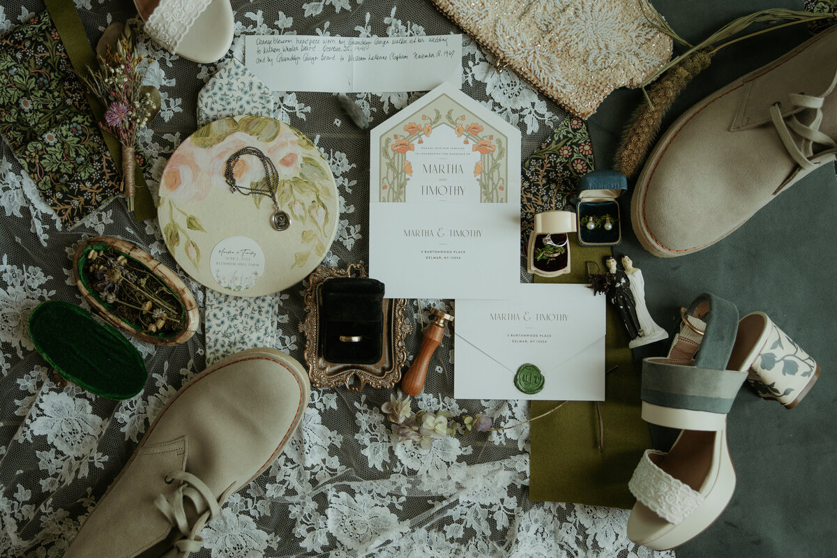 Flat lay of wedding details including shoes, invitations, jewelry, and floral decorations arranged on a lace fabric background