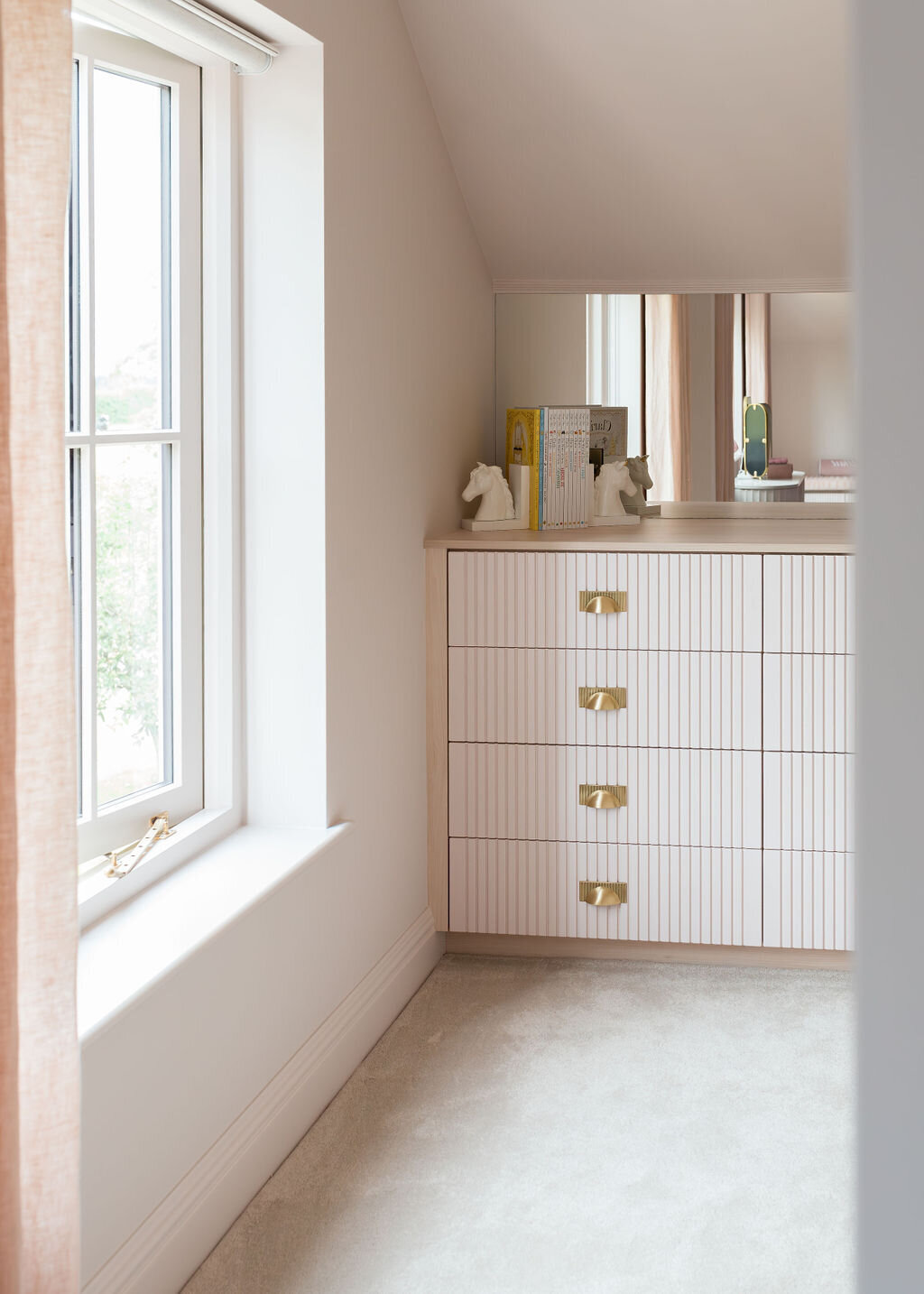 A modern pale pink dresser with gold half-moon handles sits next to a large window lettings in bright, natural light.