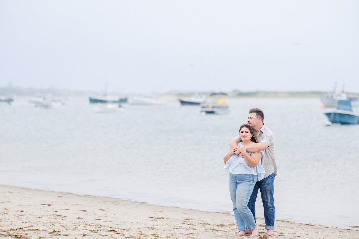 Man standing behind a woman on the beach, arms wrapped around her shoulders, both smiling at the camera with blurred boats in the background at Chatham Bars Inn in Chatham, Massachusetts — representing the portfolio of a Cape Cod engagement photographer.