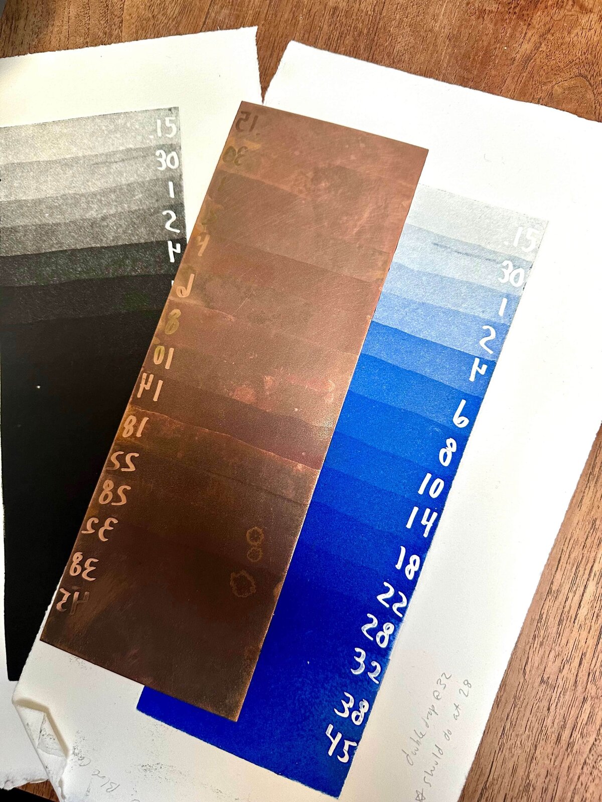 A copper timing strip is laid across two test prints—one in black and one in blue—both showing tonal steps and labeled etch times.