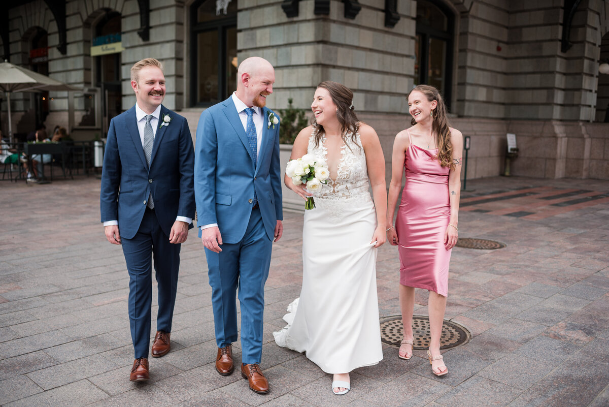 bride in a satin and lace wedding dress holding a white floral bouquet laughing with her groom as they walk down town with their bridal party captured by denver wedding photographer