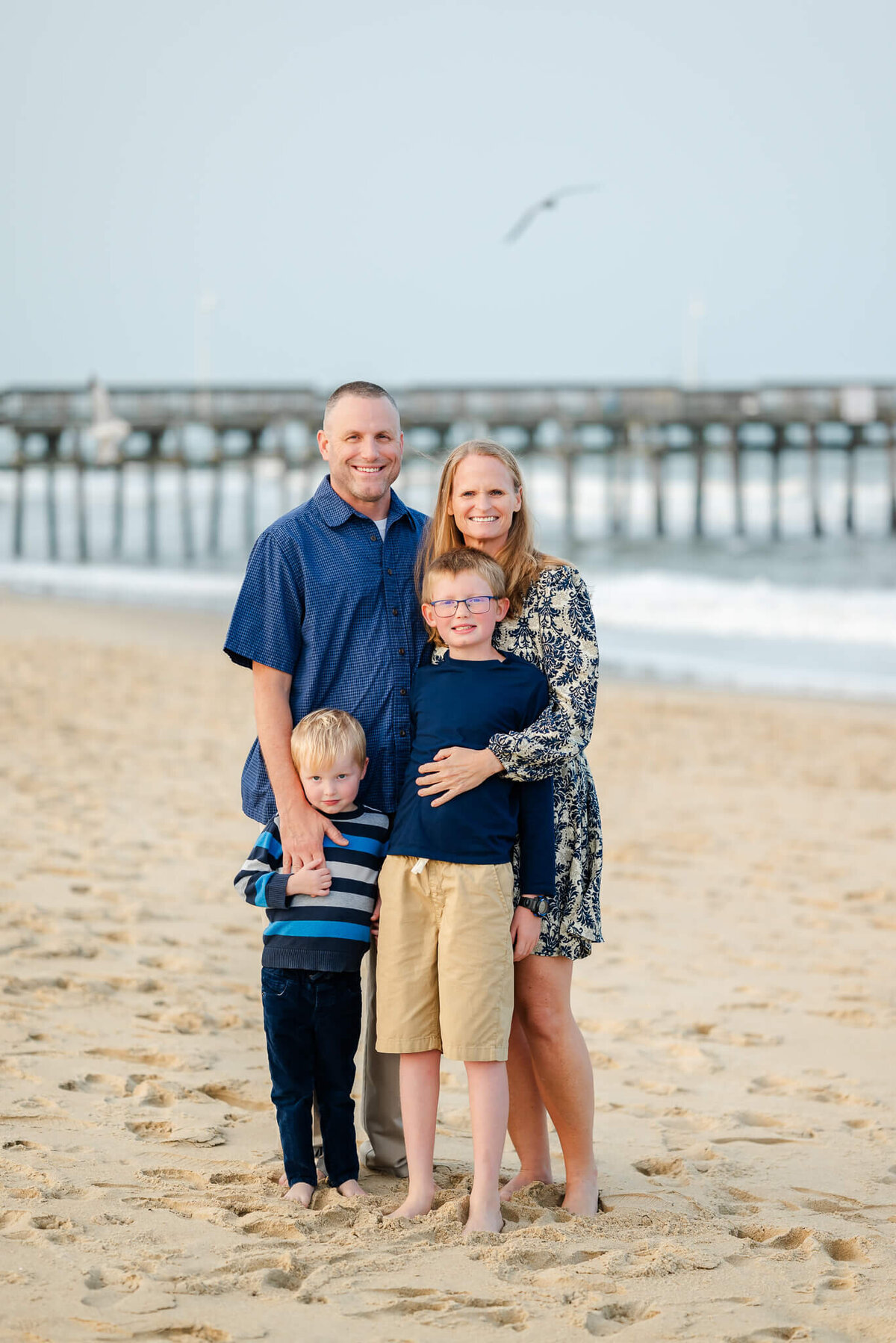 A family with a mom and dad and two young sons stands in front of a fishing pier on a beach in Hampton Roads.