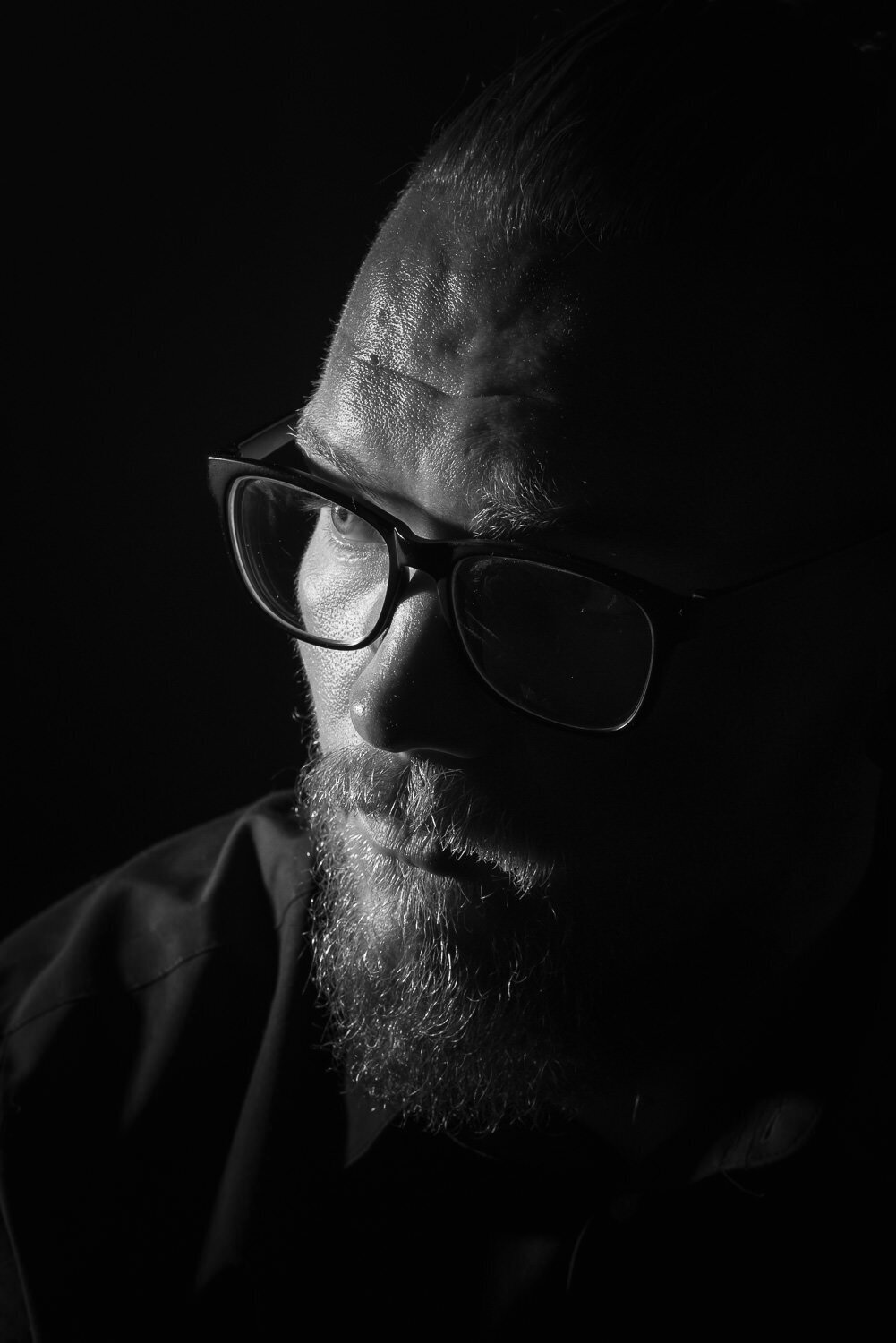 Dramatic low-key close-up portrait of a bearded man with glasses, featuring a sliver of light illuminating the edge of his face and eye for a cinematic effect.