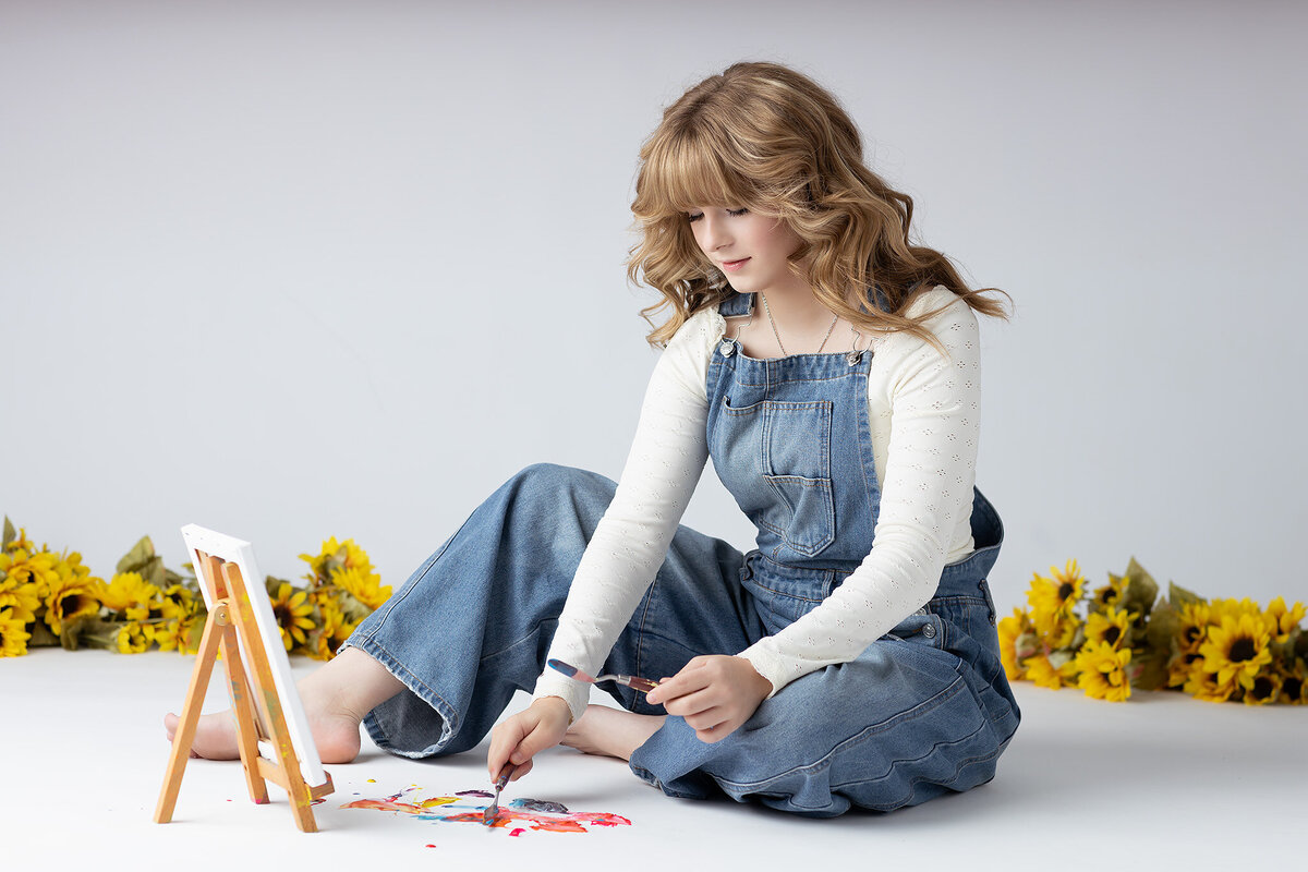 A young woman sits on the floor with her feet bare, dressed in denim overalls and a long-sleeve white top. She carefully mixes vibrant paints on the floor, surrounded by a border of sunflowers. A small easel with a blank canvas stands nearby, capturing her artistic spirit in this serene and joyful moment.