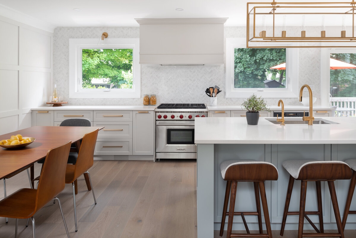 Large kitchen with white panelled feature fall, all white herringbone wall tile, white cabinets, all gold hardware and light fixtures, a large dark wood minimalistic dining room table, and an island panted soft blue complete with low-back bar stools.