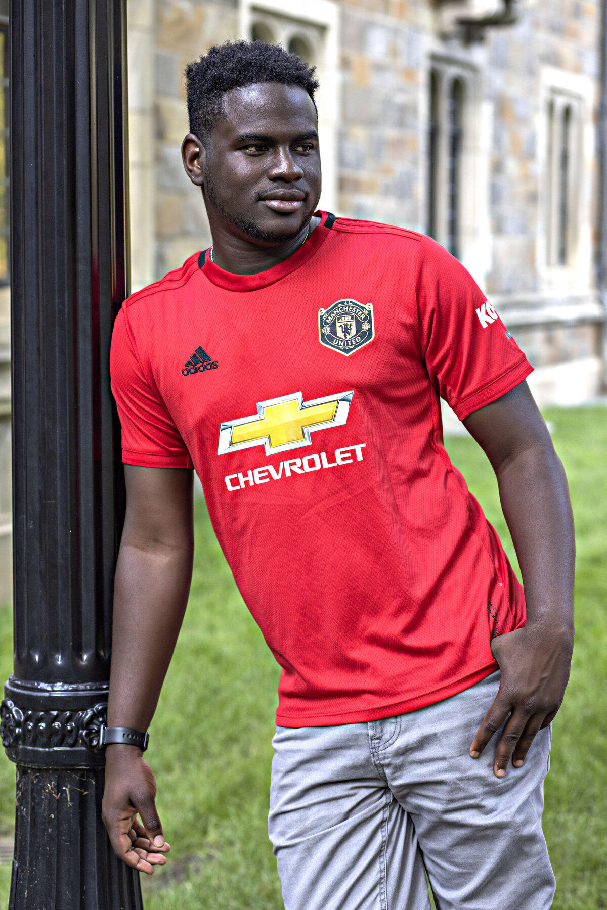 African American male wearing red sports shirt and grey jeans leaning against light pole